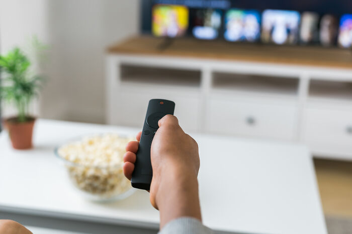 Person holding a remote control in a living room while watching TV, with a bowl of popcorn on the table, highlighting travel must-haves.