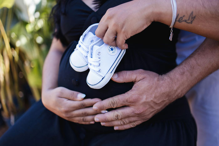 Pregnant woman holding baby shoes with returning husband’s hands on her belly.