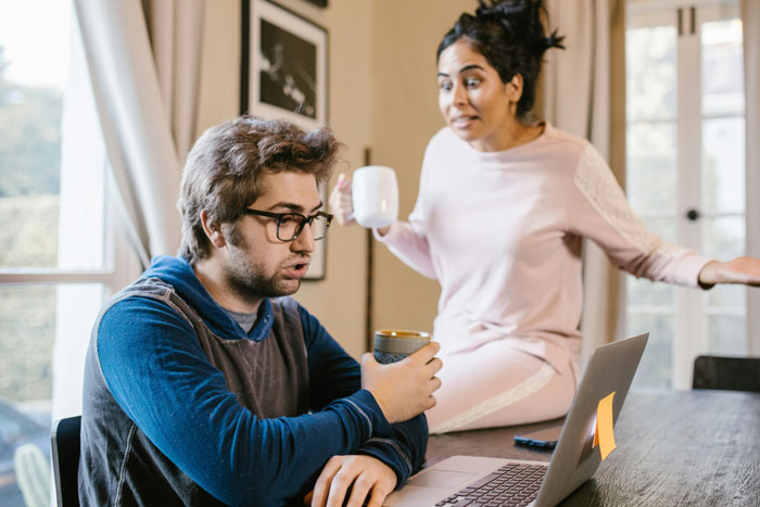 Man with glasses at laptop, woman in loungewear holding mug, discussing returning shoes.