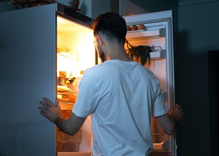 A man in a white t-shirt opening a fridge at night, searching for food.