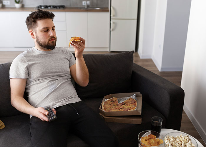 Man sitting on a couch eating pizza, surrounded by snacks, with a remote in hand.