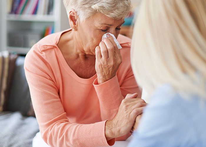 Elderly woman in an orange sweater wiping tears with tissue, comforted by another person, room conflicts theme.