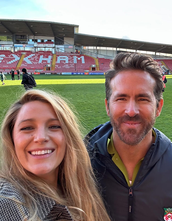 Selfie of a smiling woman with Ryan Reynolds at a stadium, both dressed casually, with empty stands in the background.