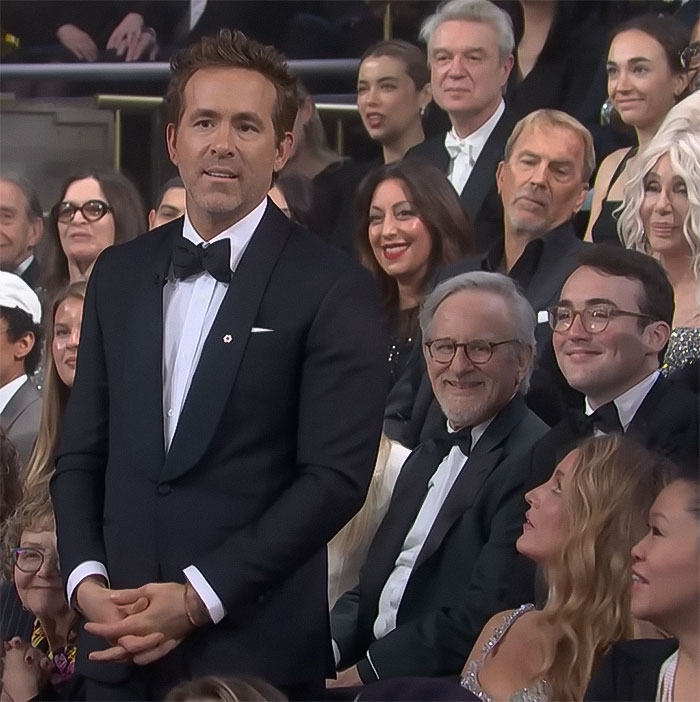 Celebrity in a tuxedo standing among a seated audience at a formal event.