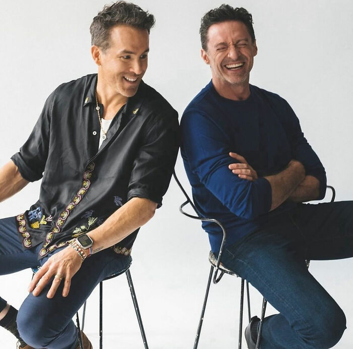 Two men laughing together while seated, wearing casual shirts and jeans, against a plain background.