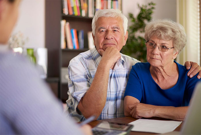 Elderly couple sitting together at a table, listening to a person across them, surrounded by bookshelves.