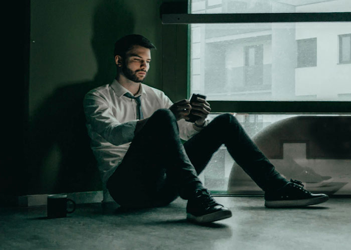 Man in a white shirt, sitting on the floor, looking at his phone with a serious expression.