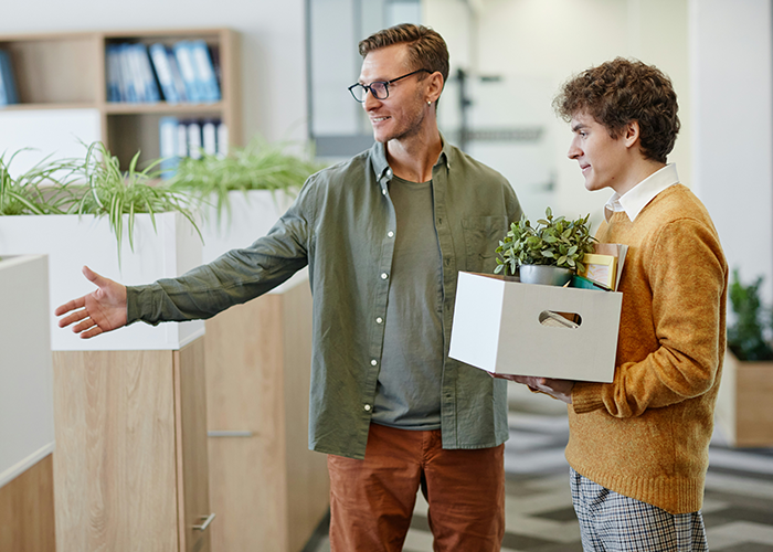 Two men in an office, one carrying a box, discussing work responsibilities and tasks.