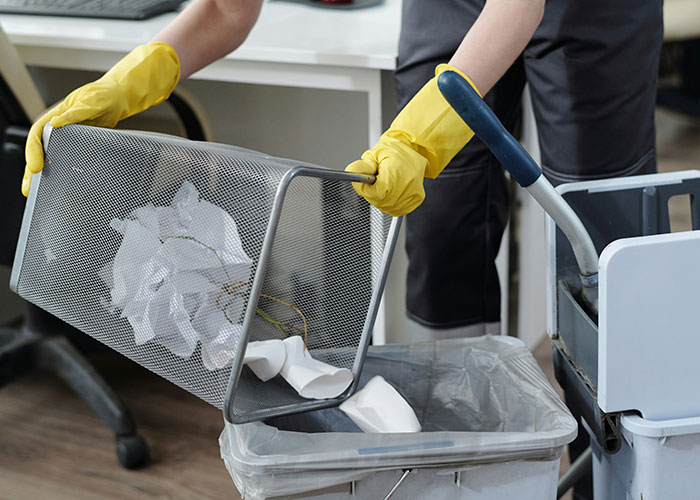 Worker cleaning office, emptying trash into bin with yellow gloves, emphasizing new guy's added workload.