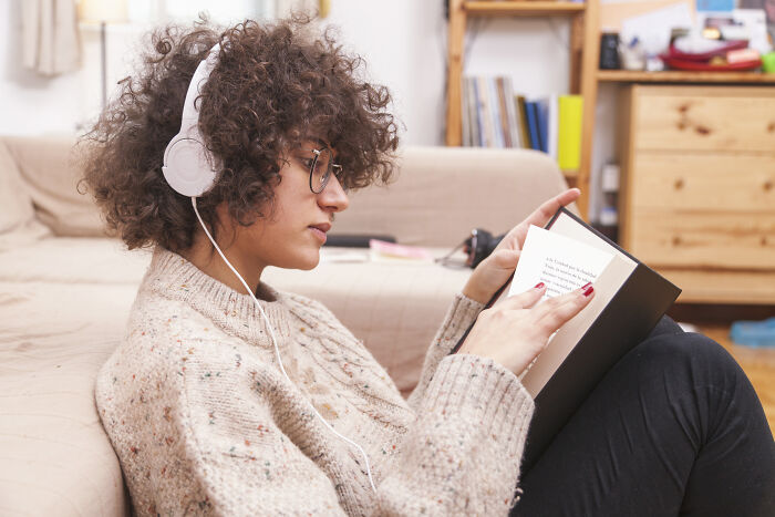 Person with curly hair and glasses listening to music and reading on a couch, highlighting travel must-haves for packing.