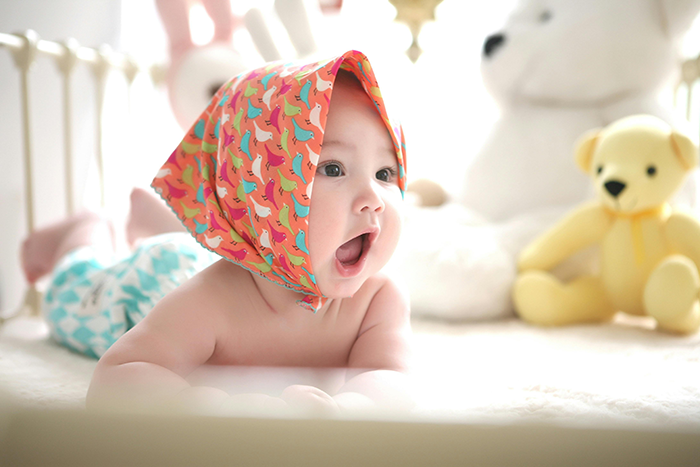 Baby with colorful scarf, lying in crib next to stuffed animals; related to babysitting and family dynamics.