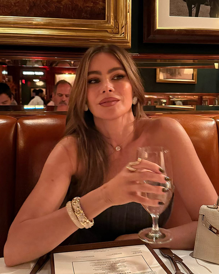 Woman seated in a restaurant holding a wine glass, wearing jewelry and a black strapless top.