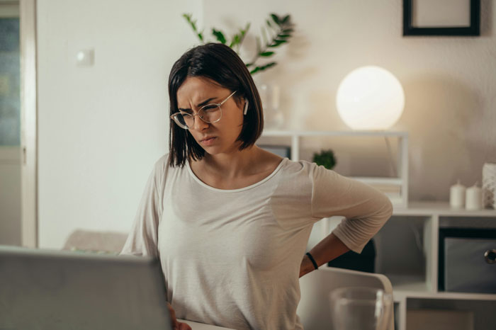 Woman with a concerned expression looking at a laptop, wearing glasses and a white shirt, in a living room setting.