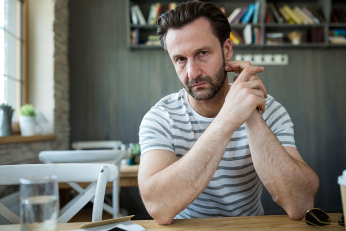 Man in striped shirt sitting pensively at a table, with bookshelf in the background, contemplating family revelations.