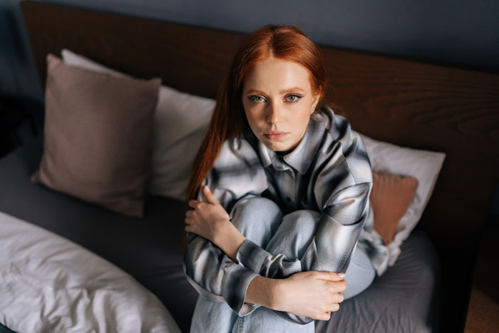 Woman sitting on a bed, looking thoughtful, wearing a plaid shirt.