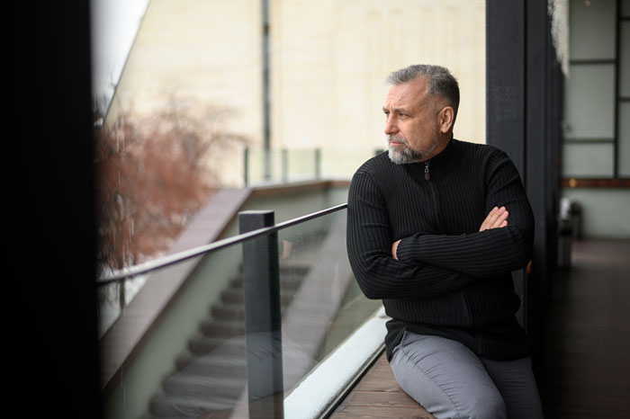 A man in a black sweater looking pensive while seated indoors, reflecting on choosing wife over daughter for wedding decision.