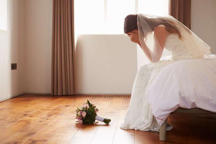 Bride sitting on a bed in distress, wedding bouquet on the floor; concept of wedding and family tension.