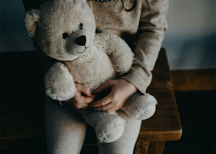 Child holding a teddy bear on a wooden bench, symbolizing strange punishment themes.