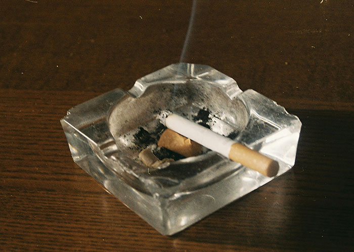 Glass ashtray with a lit cigarette on a wooden table, related to strange punishments.