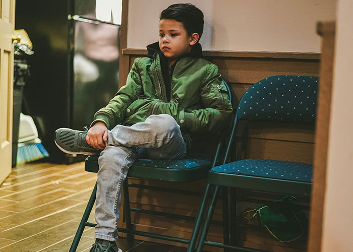 Child sitting on chairs, wearing a green jacket, appearing thoughtful about strange parental punishment.