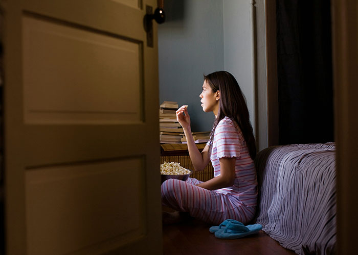 Woman in pajamas eating popcorn quietly in a dimly lit room, reflecting on strange punishment from parents.