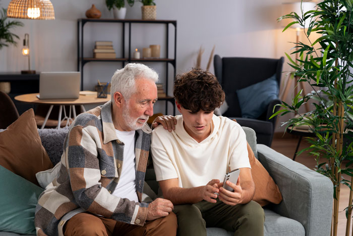 Teen showing phone to elderly man on a sofa, engaged in conversation, amidst cozy living room decor.