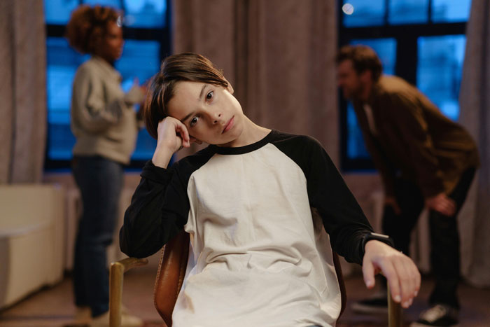 Frustrated teen in focus sitting while adults talk in the background, illustrating family neglect dynamics.