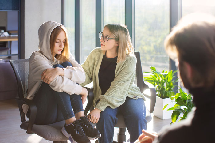 Teen expressing frustration, sitting with arms crossed, while woman offers support in a bright room with large windows.