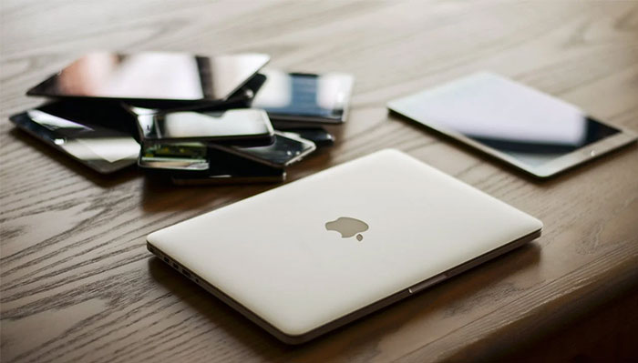 Laptop and tablets on a wooden table, highlighting corporate propaganda's impact on technology normalization.