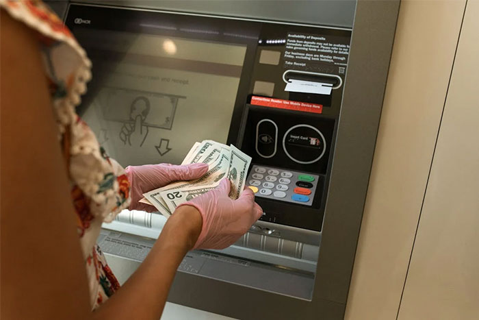 Person wearing gloves using ATM, holding cash; corporate propaganda influence on financial transactions.