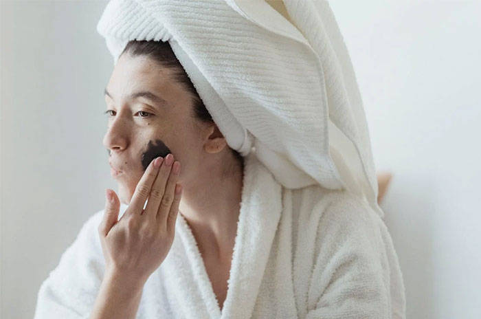 Person in a bathrobe, towel on head, applying skincare, highlighting corporate propaganda's influence on beauty routines.