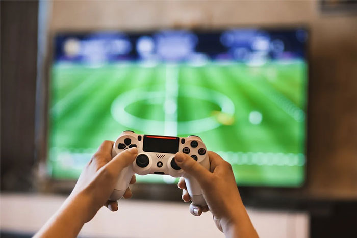 Hands holding a video game controller with a soccer game on TV; depicts corporate propaganda influence in gaming culture.