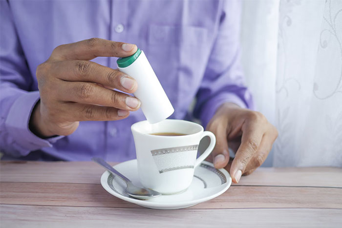 Person adding sweetener to coffee, illustrating corporate propaganda normalization in daily habits.