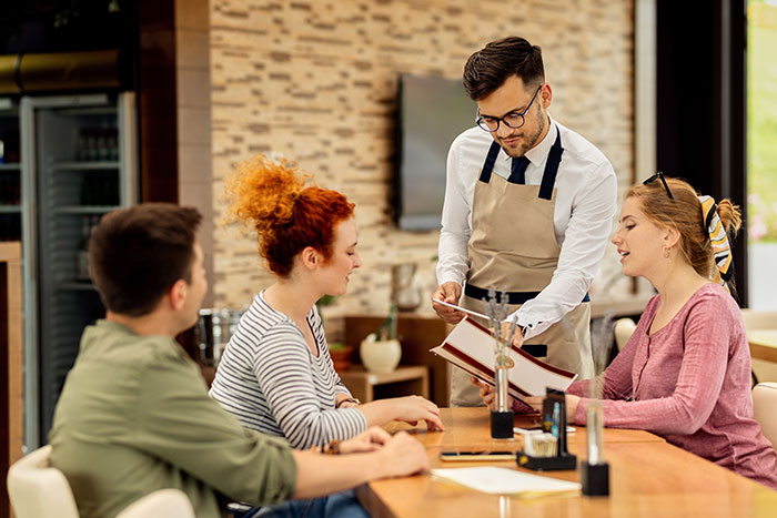 Waiter showing a menu to two women and a man in a restaurant setting.