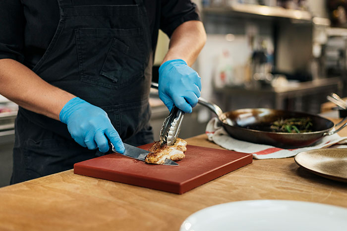 Chef wearing blue gloves, slicing chicken on a red cutting board in a restaurant kitchen environment.