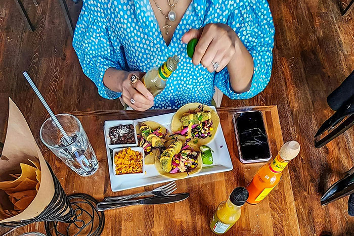 Person adding sauce to tacos at a restaurant, with drinks and a phone on the table.