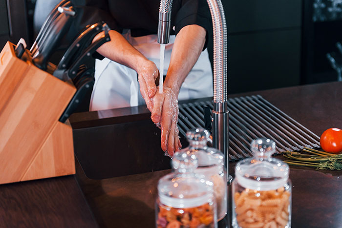 Person washing hands in a restaurant kitchen sink near knives and ingredients, suggesting hygiene awareness.