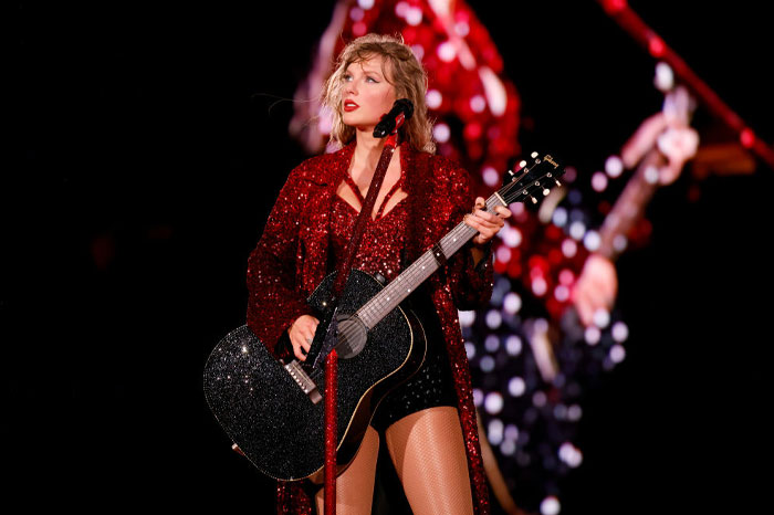 Taylor Swift performing on stage in a glittery red outfit, holding a black acoustic guitar.