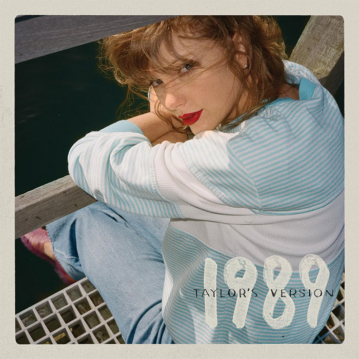 Taylor Swift sitting by water, wearing a striped sweater and jeans, featuring &lsquo;1989 Taylor&rsquo;s Version&rsquo; text.