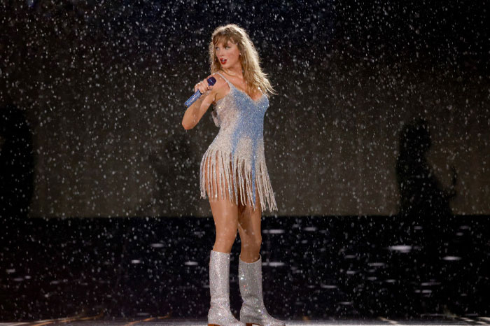 Taylor Swift performing in a sparkling outfit on stage with a microphone in hand, surrounded by a dramatic snowfall effect.