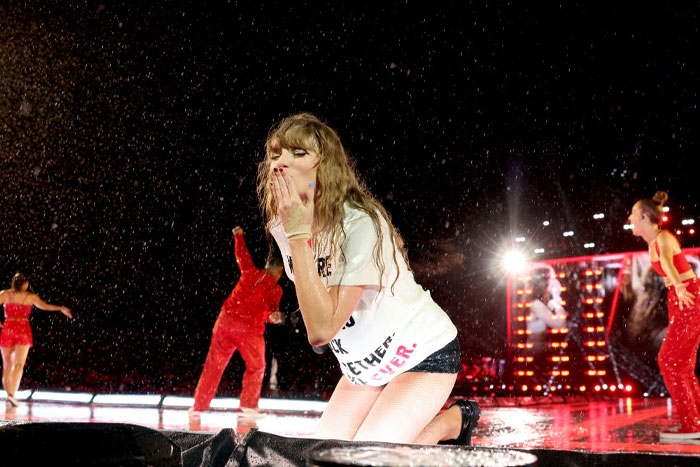 Taylor Swift performing on a wet stage, blowing a kiss, surrounded by dancers in red outfits.