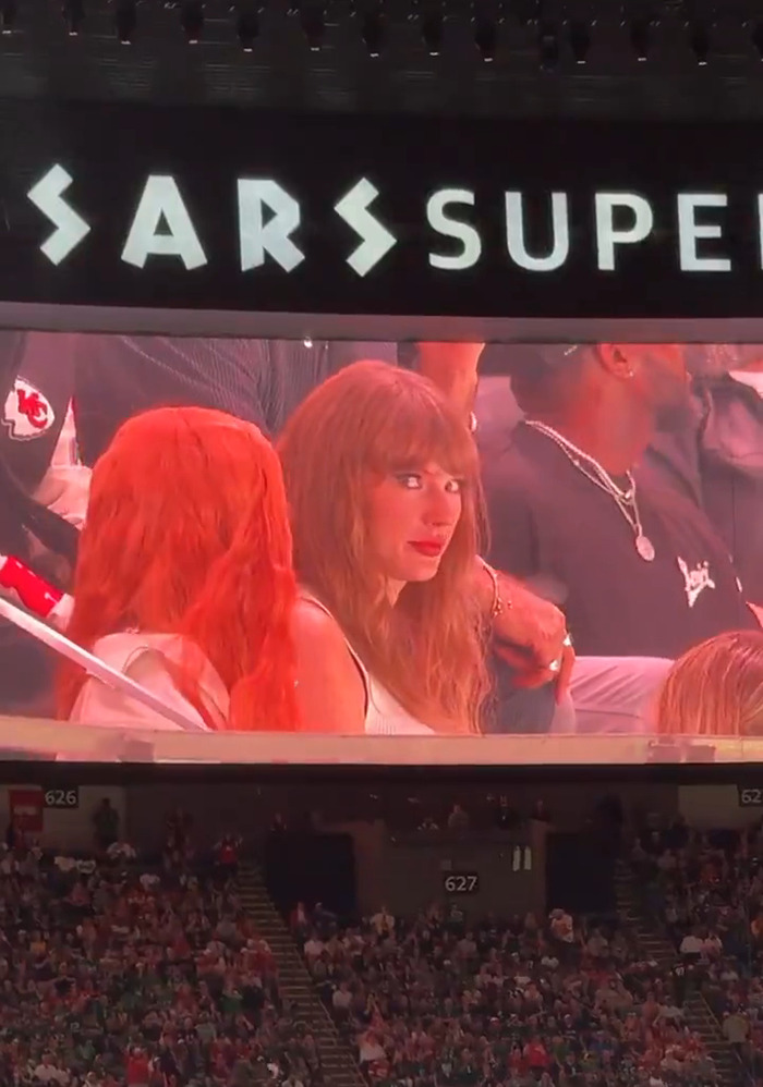 Taylor Swift reacting at the Super Bowl, her image displayed on a large screen above the audience.