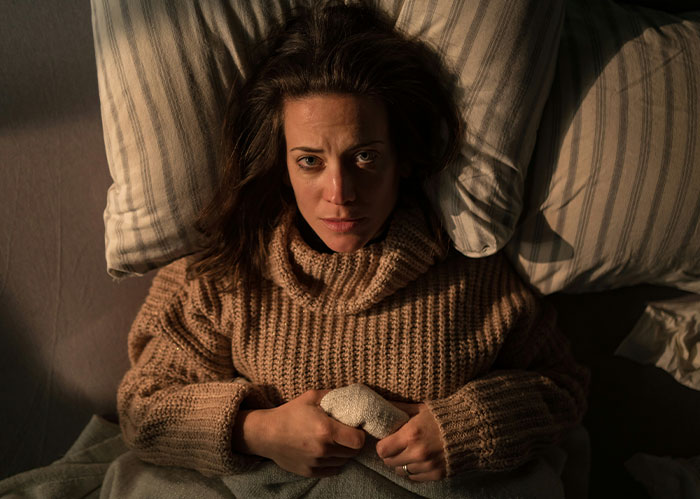 A woman lying in bed, looking concerned, wearing a brown sweater, surrounded by pillows.