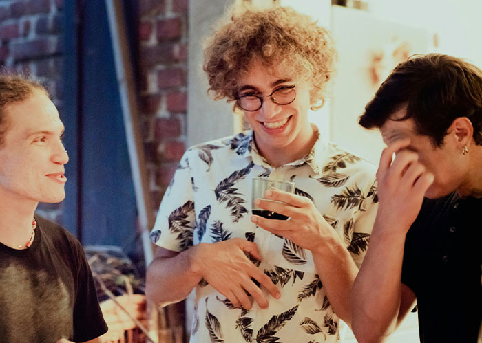 Three people laughing together in a cozy room, wearing casual clothes and holding drinks.