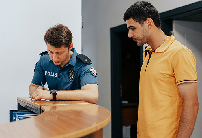 A person in a yellow shirt talks to a police officer writing at a desk, discussing a student's creepy comment.