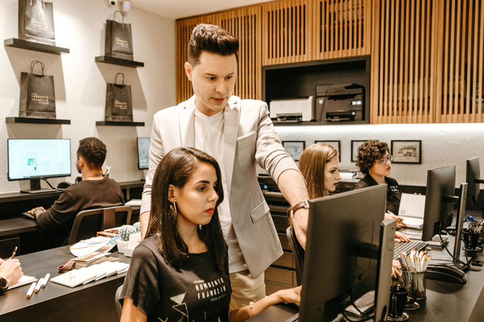 Boss trying to optimize company's structure, standing over employee at desk in a modern office setting.