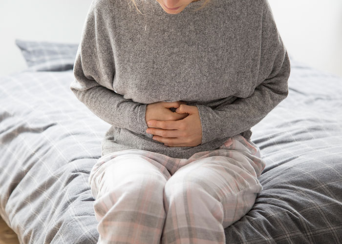 Woman sitting on a bed, wearing a gray sweater and pajama pants, holding her stomach, symbolizing things men learn about women.