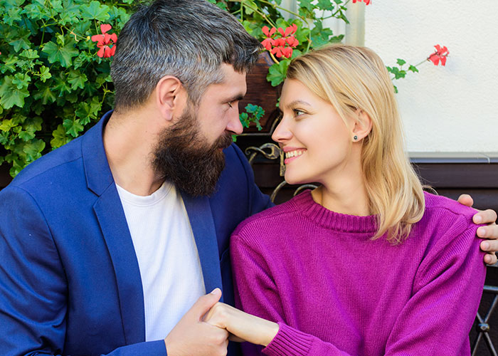 Man and woman smiling at each other, sitting on a bench surrounded by flowers.