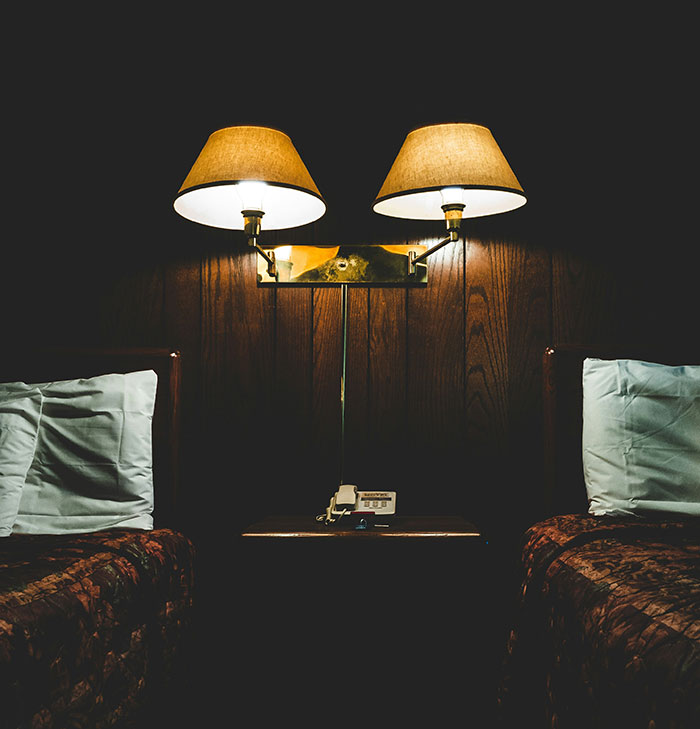 Frugal hotel room with two twin beds, vintage decor, and dual lamps above nightstand.