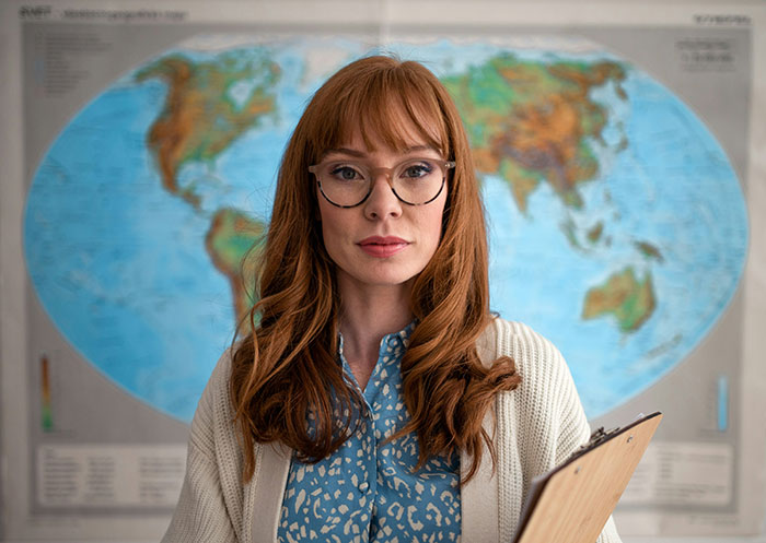 A woman in glasses holding a clipboard, standing in front of a world map, highlighting frugality decisions.